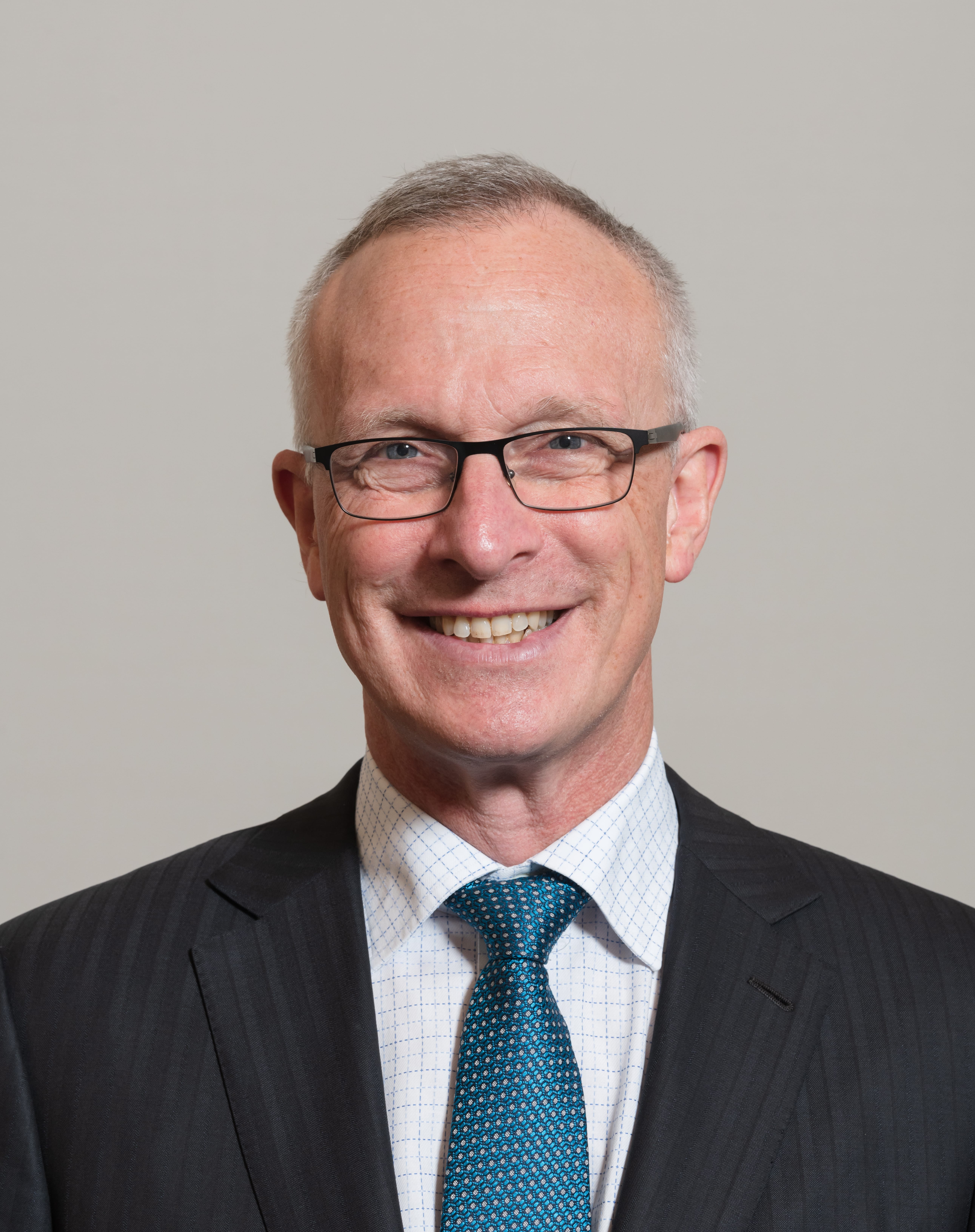 Smiling man with glasses in a dark suit and blue patterned tie.